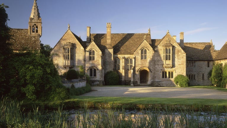 A view across the moat of the front and forecourt of Great Chalfield Manor, a medieval manor house in Wiltshire, with blue sky and low sun shining on the warm stone.